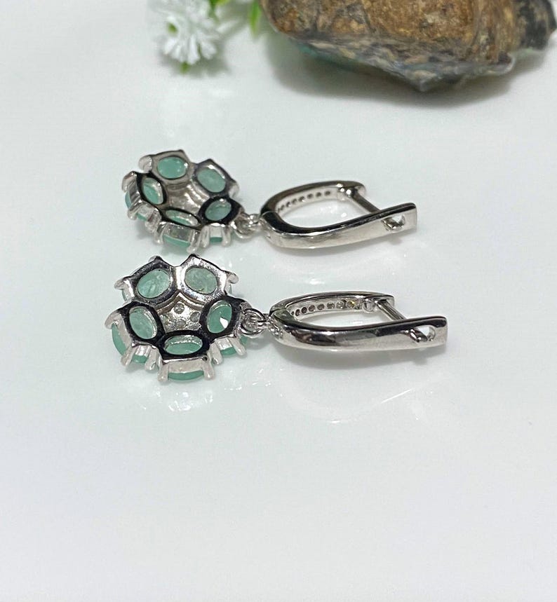 May include: A pair of silver-toned drop earrings. Each earring features a cluster of oval-shaped, light green gemstones set in a floral design. The earrings have a lever-back closure and are displayed on a white surface.