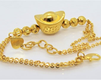 Puede incluir: Un brazalete de oro con un dije de oro en forma de lingote de oro chino. El dije tiene el número 888 en relieve. El brazalete está hecho de una cadena delicada con pequeñas cuentas redondas.