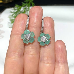 May include: A pair of silver-toned earrings featuring a floral design. Each earring has six oval-shaped, light green gemstones surrounding a cluster of smaller, clear gemstones. The earrings are held by a person's hand.