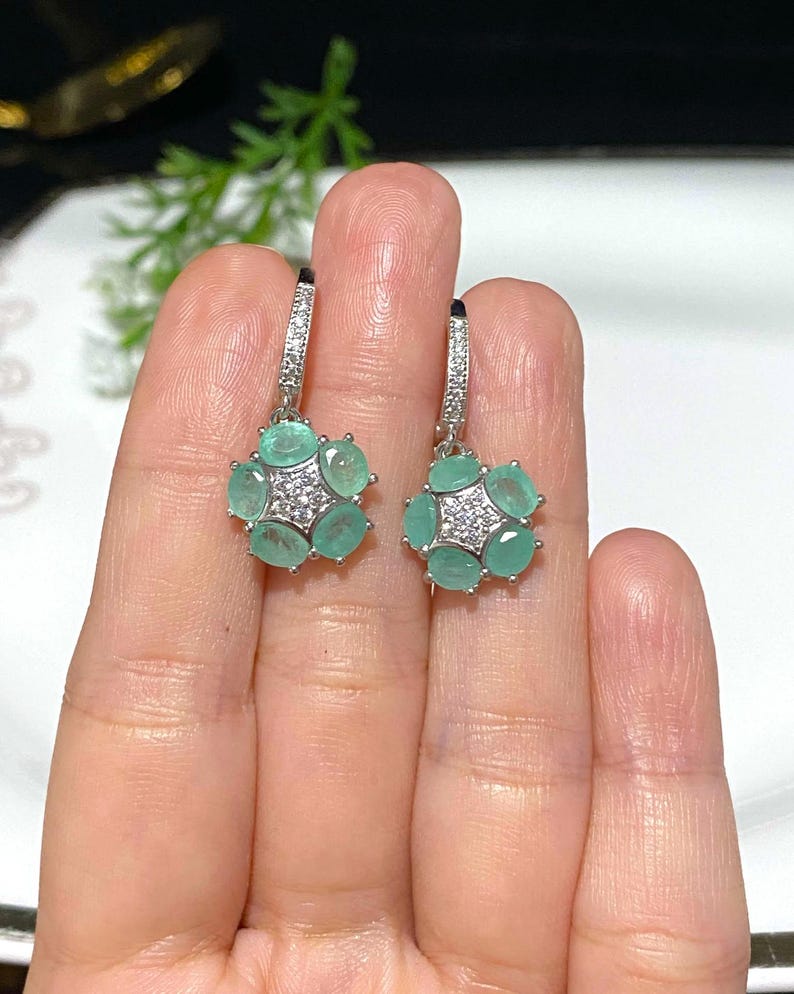 May include: A pair of silver-toned earrings featuring a floral design. Each earring has six oval-shaped, light green gemstones surrounding a cluster of smaller, clear gemstones. The earrings are held by a person's hand.