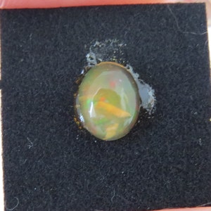 Puede incluir: Una piedra preciosa de ópalo redonda de forma ovalada con un brillo iridiscente verde, amarillo y naranja. El ópalo está colocado en una caja de exposición de terciopelo negro.