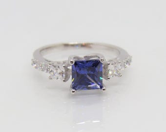 Op de afbeelding: Een zilveren ring met een vierkante blauwe edelsteen omringd door kleinere heldere edelstenen.