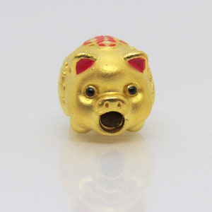 May include: A small, gold-colored piggy bank charm with red ears and a black nose. The piggy bank has a small opening on the top for coins.