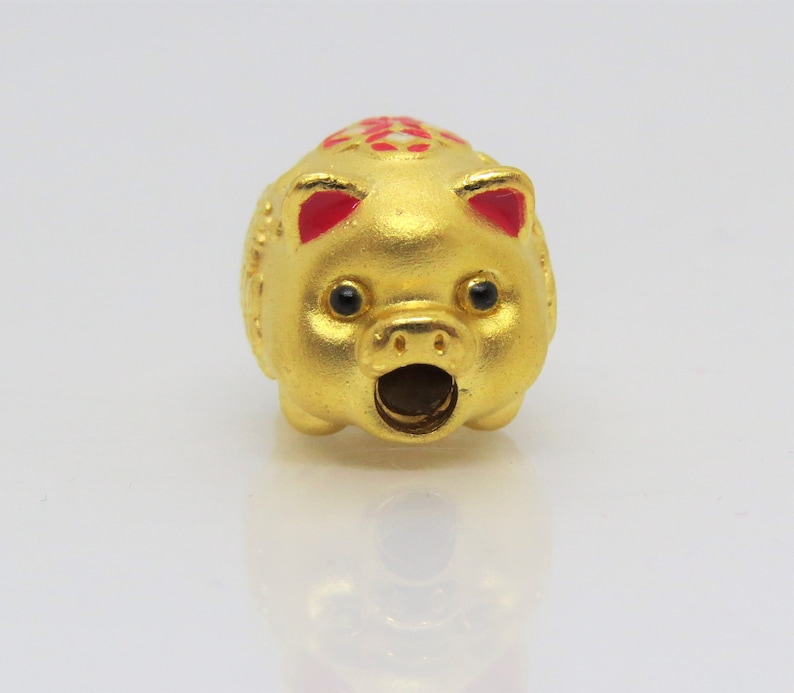 May include: A small, gold-colored piggy bank charm with red ears and a black nose. The piggy bank has a small opening on the top for coins.