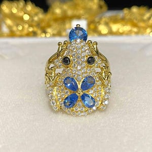 Könnte beinhalten: Ein goldfarbener Ring mit einem Frosch-Design, besetzt mit blauen und weißen Edelsteinen. Der Ring hat einen großen blauen Stein oben, zwei schwarze Steine als Augen und eine Ansammlung kleinerer weißer und blauer Steine. Der Ring ist vor einem weißen Hintergrund platziert.