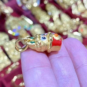 May include: A gold-colored pendant with a cartoon-like design, featuring blue, red, and green accents. The pendant has a loop for a chain and is held in a person's hand. The background is blurred with other gold-colored items.