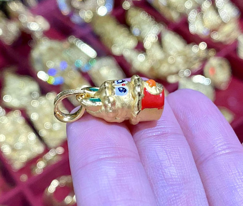 May include: A gold-colored pendant with a cartoon-like design, featuring blue, red, and green accents. The pendant has a loop for a chain and is held in a person's hand. The background is blurred with other gold-colored items.