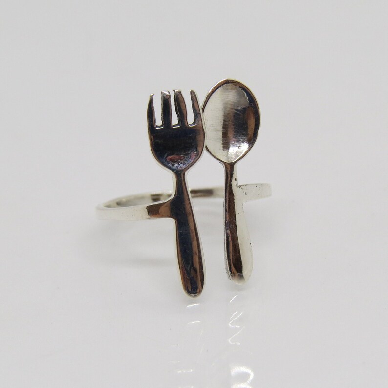 Puede incluir: Un anillo de plata con un dise&ntilde;o de tenedor y cuchara. El tenedor y la cuchara est&aacute;n conectados en la base del anillo.