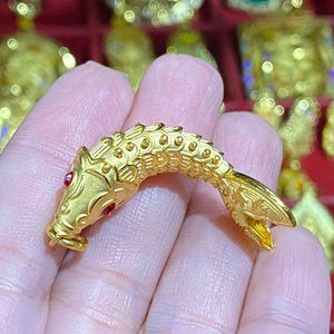 Puede incluir: Un amuleto de pez de color dorado con ojos rojos. El amuleto est&aacute; detallado con escamas y aletas.