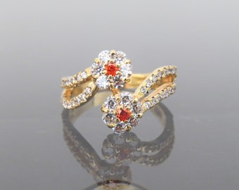 May include: A gold ring with two flower-shaped settings. Each setting has a cluster of clear cubic zirconia stones surrounding a single red stone.