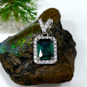 Puede incluir: Un colgante de plata con una gran piedra preciosa verde esmeralda rodeada por un halo de piedras preciosas más pequeñas y transparentes.