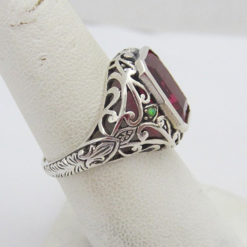 May include: A silver ring with an ornate filigree design features a large, rectangular, red gemstone and a smaller, green gemstone.