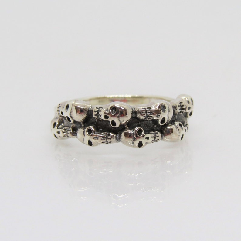 May include: A silver ring with a band of skulls. The skulls are arranged in a staggered pattern, with some facing forward and others facing sideways.