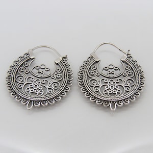 May include: A pair of silver-colored hoop earrings with an intricate, openwork design. The earrings feature a circular shape with detailed floral and scroll patterns, creating a delicate and ornate appearance. The metal has a polished finish.