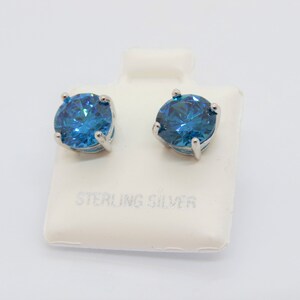 Puede incluir: Un par de pendientes de plata de ley con piedras redondas azules. Los pendientes se muestran en una tarjeta blanca con el texto "Sterling Silver".