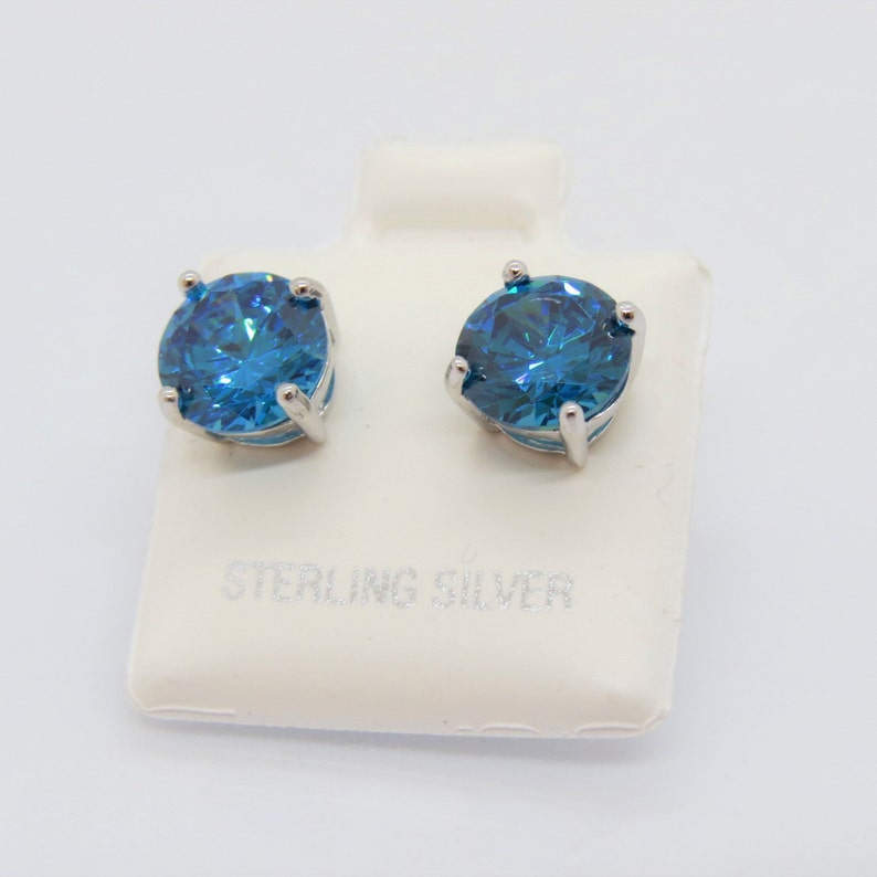 Puede incluir: Un par de pendientes de plata de ley con piedras redondas azules. Los pendientes se muestran en una tarjeta blanca con el texto "Sterling Silver".