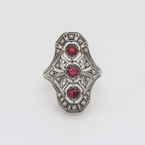 May include: A silver ring with a filigree design and three red gemstones. The ring is set with small white pearls.