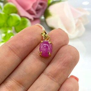 Puede incluir: Un colgante de piedra ovalada rosa con engaste de oro. El colgante se sostiene en una mano.
