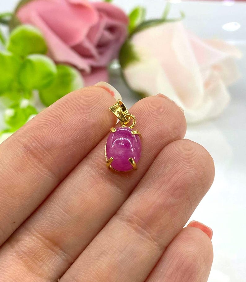 Puede incluir: Un colgante de piedra ovalada rosa con engaste de oro. El colgante se sostiene en una mano.