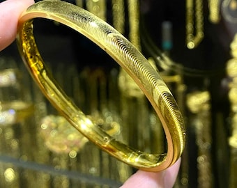 May include: A gold bangle bracelet with a textured surface. The bracelet is a bright, shiny gold color and appears to be made of metal. The design features a series of curved lines along the outer edge.