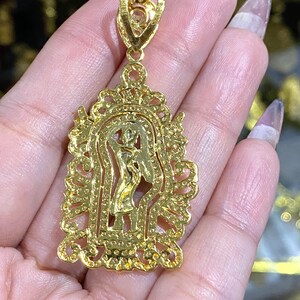 May include: A gold-colored pendant featuring a detailed religious figure within an ornate, filigree-style frame. The pendant has a bail for a chain and is likely made of gold or gold-plated metal. The design includes small, sparkling accents.