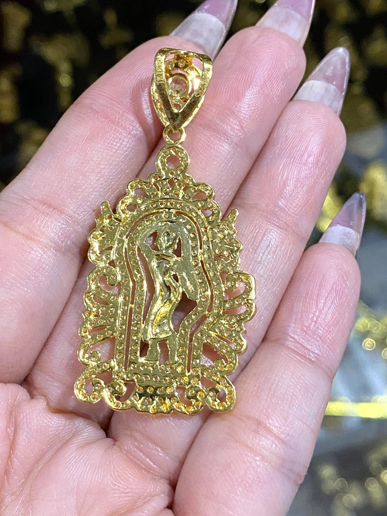 May include: A gold-colored pendant featuring a detailed religious figure within an ornate, filigree-style frame. The pendant has a bail for a chain and is likely made of gold or gold-plated metal. The design includes small, sparkling accents.