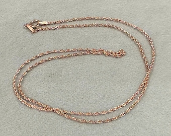 May include: A delicate rose gold necklace with a fine, twisted chain. The necklace is laid out on a light grey surface, showcasing its intricate design and the warm hue of the metal. The clasp is visible at the top.