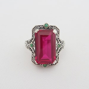 May include: A silver ring with a large, rectangular, pink gemstone surrounded by a filigree design and four smaller green gemstones.