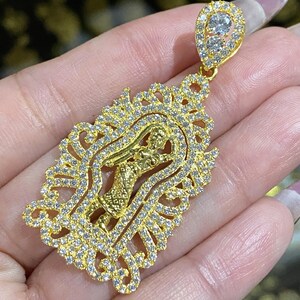 May include: A gold-colored pendant with a detailed design. The pendant features a central figure within an ornate, filigree-style frame, encrusted with small, clear gemstones. The top of the pendant has a teardrop-shaped bail, also set with gemstones.