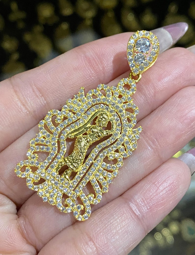 May include: A gold-colored pendant with a detailed design. The pendant features a central figure within an ornate, filigree-style frame, encrusted with small, clear gemstones. The top of the pendant has a teardrop-shaped bail, also set with gemstones.