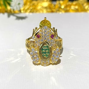 May include: A gold ring with a frog design. The frog is adorned with white, green, and pink stones.