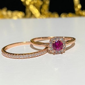 May include: A set of two rose gold rings with a ruby and diamond accents. The larger ring features a ruby center stone surrounded by a halo of diamonds. The smaller ring is a band with diamonds set in a pave setting.