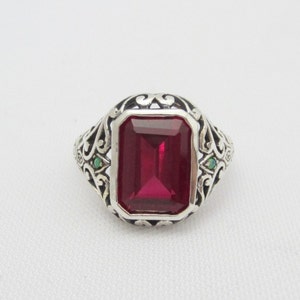 May include: A silver ring with an emerald-cut red gemstone surrounded by an ornate silver filigree design. Two smaller green gemstones are set on either side of the main stone.