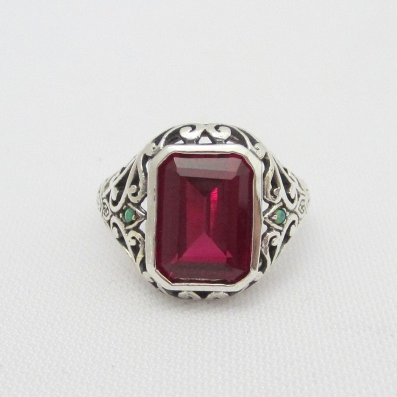 May include: A silver ring with an emerald-cut red gemstone surrounded by an ornate silver filigree design. Two smaller green gemstones are set on either side of the main stone.