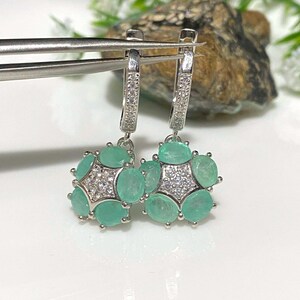 May include: A pair of silver earrings with a cluster of oval-shaped, light green gemstones and small, clear stones. The earrings are held by silver tweezers against a blurred background.