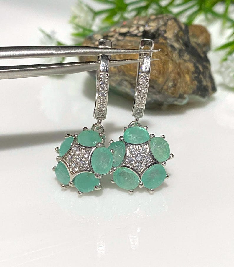 May include: A pair of silver earrings with a cluster of oval-shaped, light green gemstones and small, clear stones. The earrings are held by silver tweezers against a blurred background.