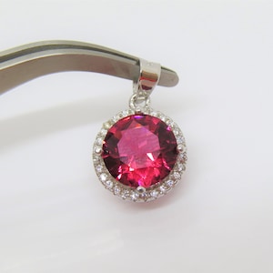 Peut inclure: Un pendentif en argent avec une grande pierre précieuse rouge ronde entourée d'un halo de petites pierres précieuses claires.