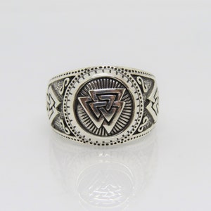 May include: A silver ring with a detailed design featuring a stylised triangular symbol in the centre. The ring has a textured surface and a circular band.