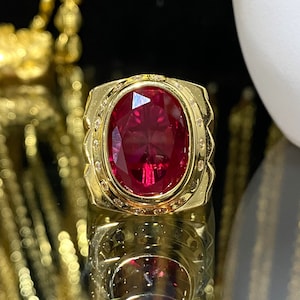 Könnte beinhalten: Ein goldener Ring mit einem großen, ovalen, roten Edelstein, der von kleineren, weißen Edelsteinen umgeben ist.