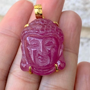 May include: A pink ruby gemstone carved into the shape of a Buddha's face, set in a gold pendant.