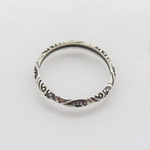 May include: A silver band ring with a delicate, intricate design. The band is thin and features a repeating pattern of swirls and dots.