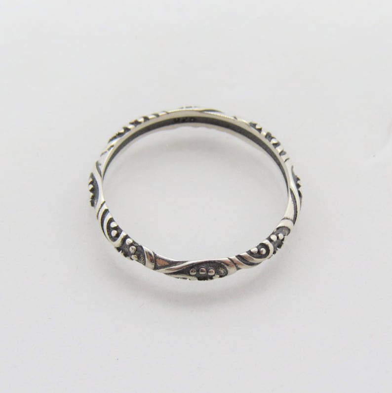 May include: A silver band ring with a delicate, intricate design. The band is thin and features a repeating pattern of swirls and dots.