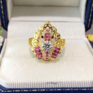 May include: A gold-colored ring featuring a central, round, clear gemstone surrounded by smaller, pink gemstones. The ring has a detailed, ornate design with additional small, clear gemstones and black accents.
