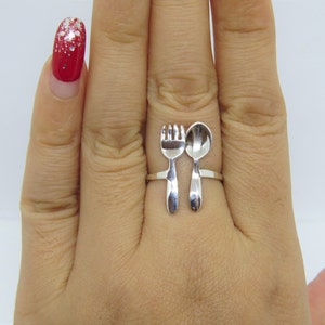 Puede incluir: Un anillo de plata con un dise&ntilde;o de tenedor y cuchara. El anillo se lleva en un dedo con esmalte de u&ntilde;as rojo.