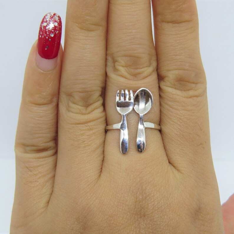Puede incluir: Un anillo de plata con un dise&ntilde;o de tenedor y cuchara. El anillo se lleva en un dedo con esmalte de u&ntilde;as rojo.