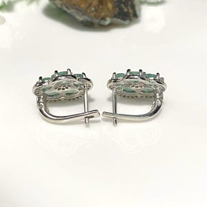 May include: A pair of silver-toned earrings with a floral design. Each earring features a cluster of oval-shaped, light green gemstones set within a flower-shaped frame. The earrings have a lever-back closure.