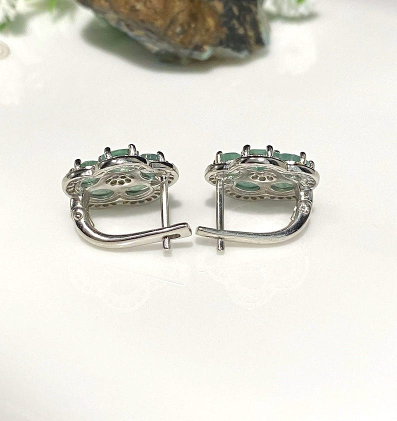 May include: A pair of silver-toned earrings with a floral design. Each earring features a cluster of oval-shaped, light green gemstones set within a flower-shaped frame. The earrings have a lever-back closure.