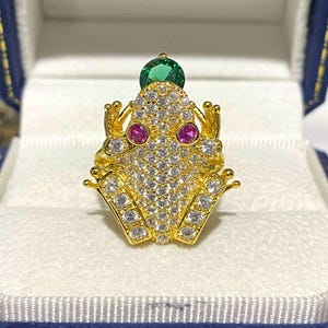以下が含まれることがあります： クリアとピンクの宝石が付いた、金色のカエルリング。カエルの目はピンク色で、頭には緑色の宝石が付いています。リングは青と白の箱に入っています。