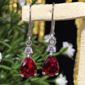 May include: A pair of silver dangle earrings featuring a teardrop-shaped red gemstone with a smaller clear gemstone above it.