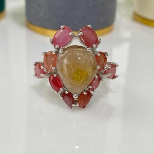 May include: A silver ring featuring a large, teardrop-shaped, yellow-brown gemstone at the center, surrounded by smaller, oval and teardrop-shaped red and orange gemstones. The ring's design is intricate and eye-catching, showcasing a floral or starburst pattern.